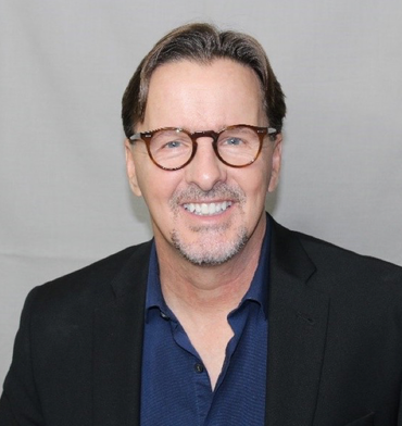 Man with glasses smiles, wearing a dark blazer and blue shirt.