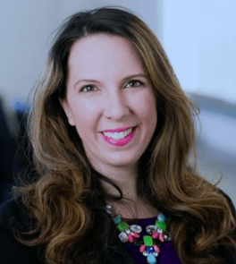 Woman with long, wavy brown hair smiles at the camera, wearing a black jacket and colorful necklace.
