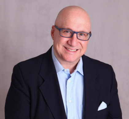 Bald man with glasses wearing a blue shirt, navy blazer, and smiling. Gray background.