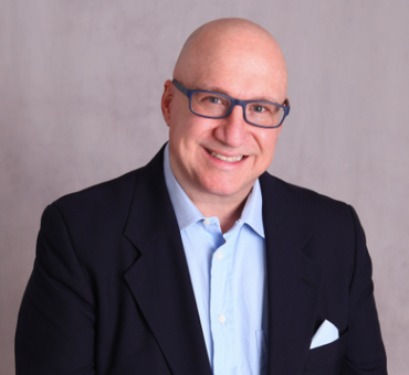 Bald man with glasses wearing a blue shirt, navy blazer, and smiling. Gray background.