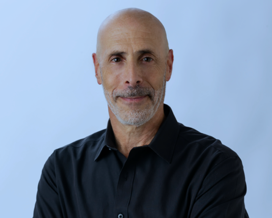 Bald man with a graying goatee in a black shirt, smiling slightly, against a light blue background.