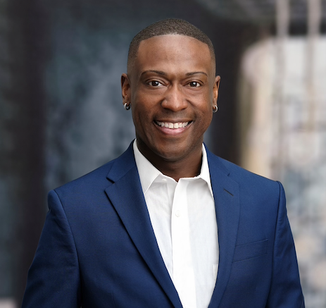 Man in a blue suit smiling, wearing small hoop earrings. Blurred city background.
