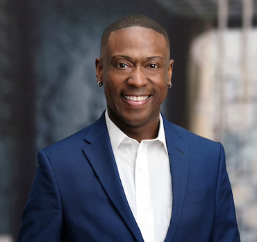 Man in a blue suit smiling, wearing small hoop earrings. Blurred city background.