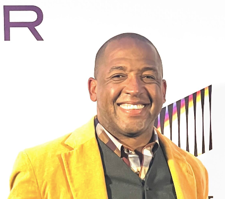 Man smiling, wearing yellow jacket over dark vest and patterned shirt, white backdrop.