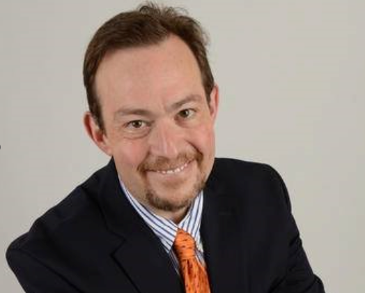 Man in a suit smiles at the camera, with a gold tie and striped shirt.