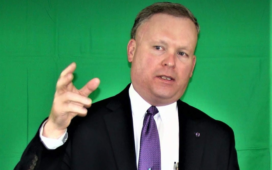 Man in suit gesturing with hand, speaking in front of a green backdrop.