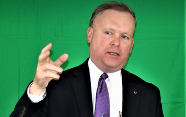 Man in suit gesturing with hand, speaking in front of a green backdrop.