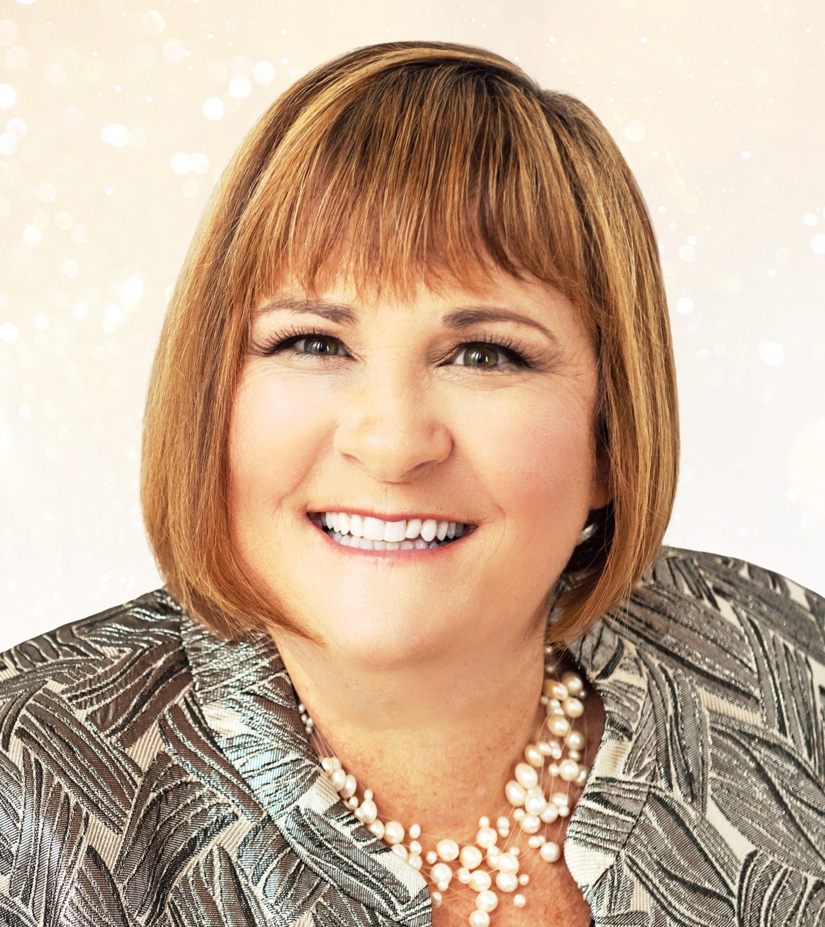 Woman with a bob hairstyle, wearing a patterned jacket and pearl necklace, smiling.