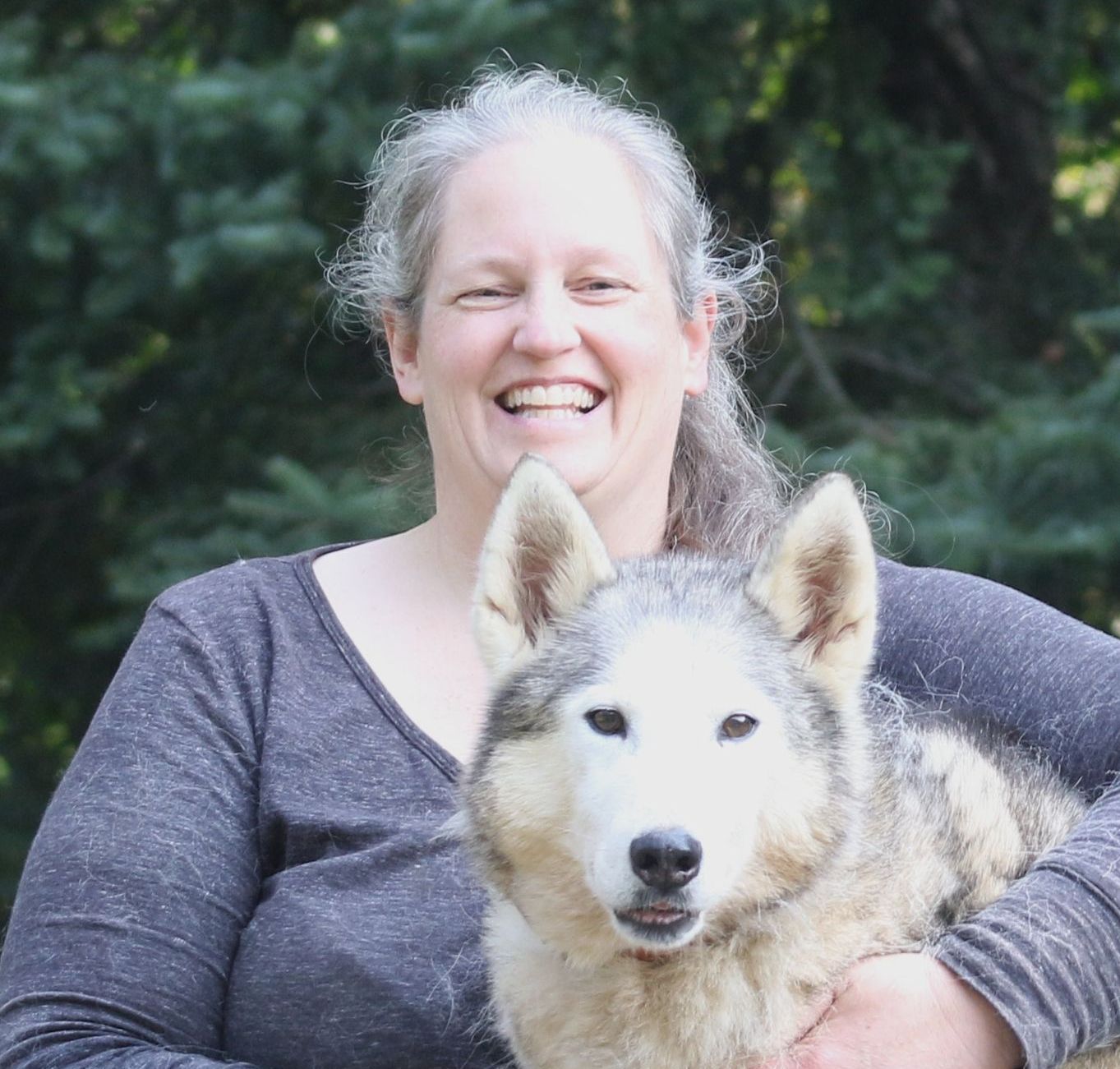 Woman smiles, holding a light-colored dog. They are outside with green trees in the background.