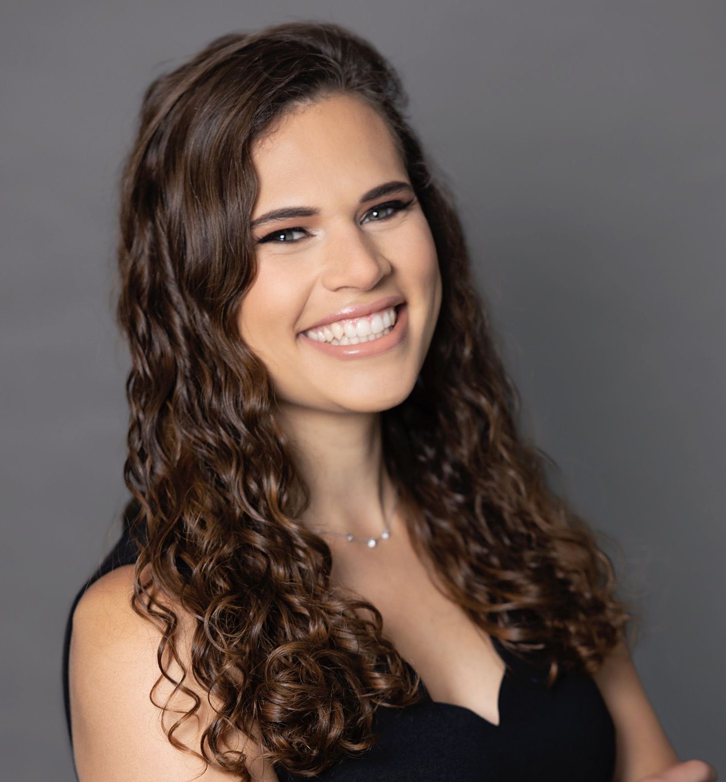 Smiling woman with curly brown hair, wearing a black top, against a gray background.