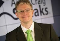 Man with glasses, wearing a suit and green tie, smiling at the camera, standing in front of a backdrop with text.