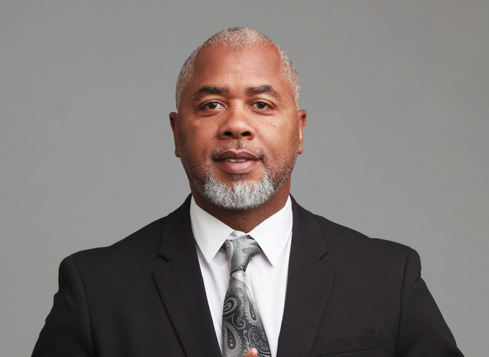 Man in a black suit with a patterned tie; he has gray hair and beard, and is looking at the camera.