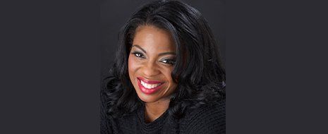 Woman with dark hair smiles, wearing dark clothing against a dark background.