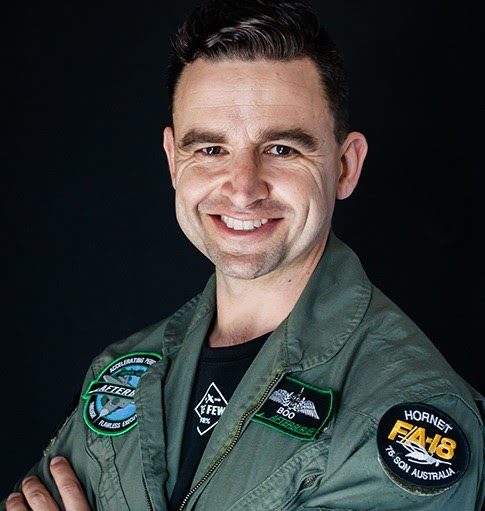 Man wearing a green flight suit smiles with arms crossed. Patches on suit: 