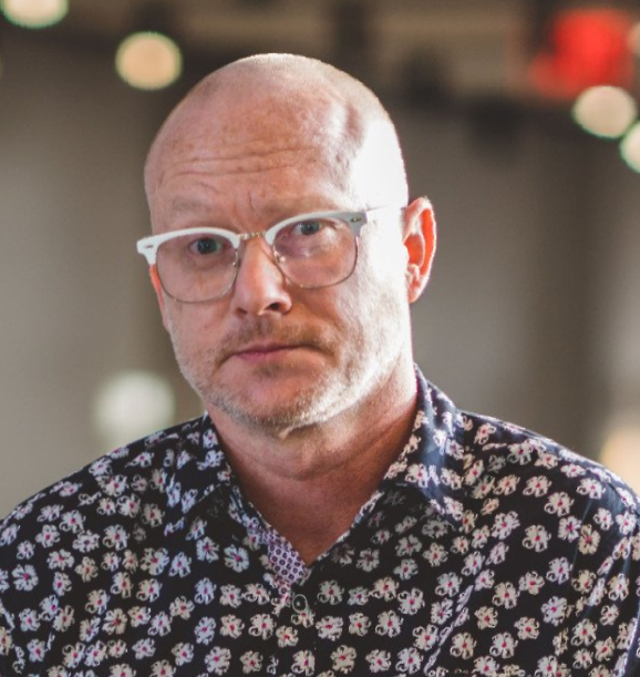 Bald man wearing glasses, patterned shirt, looking directly at the viewer.