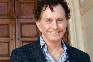 Man with curly hair smiling, wearing a blue blazer and open-collared shirt, set against a wooden door.