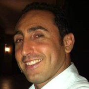 Man smiles, illuminated by a warm light; wearing a white collared shirt, dark hair.