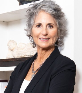 Woman with gray curly hair, wearing a black blazer and necklace, smiles at the camera, standing in front of a shelf.