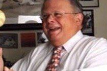Man wearing glasses, white shirt, and patterned tie, smiling and looking to the side.