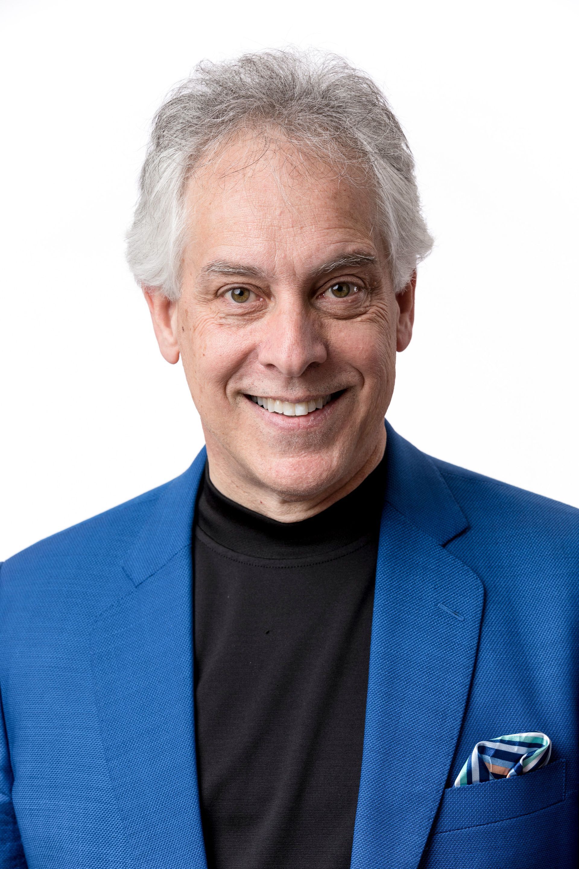 Man with gray hair, smiling, wearing a blue jacket and black shirt, pocket square visible.