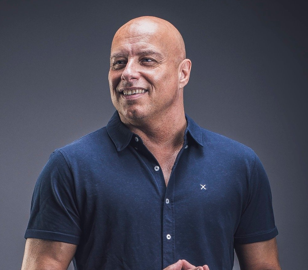 Bald man with a slight smile wearing a blue polo shirt, against a gray backdrop.