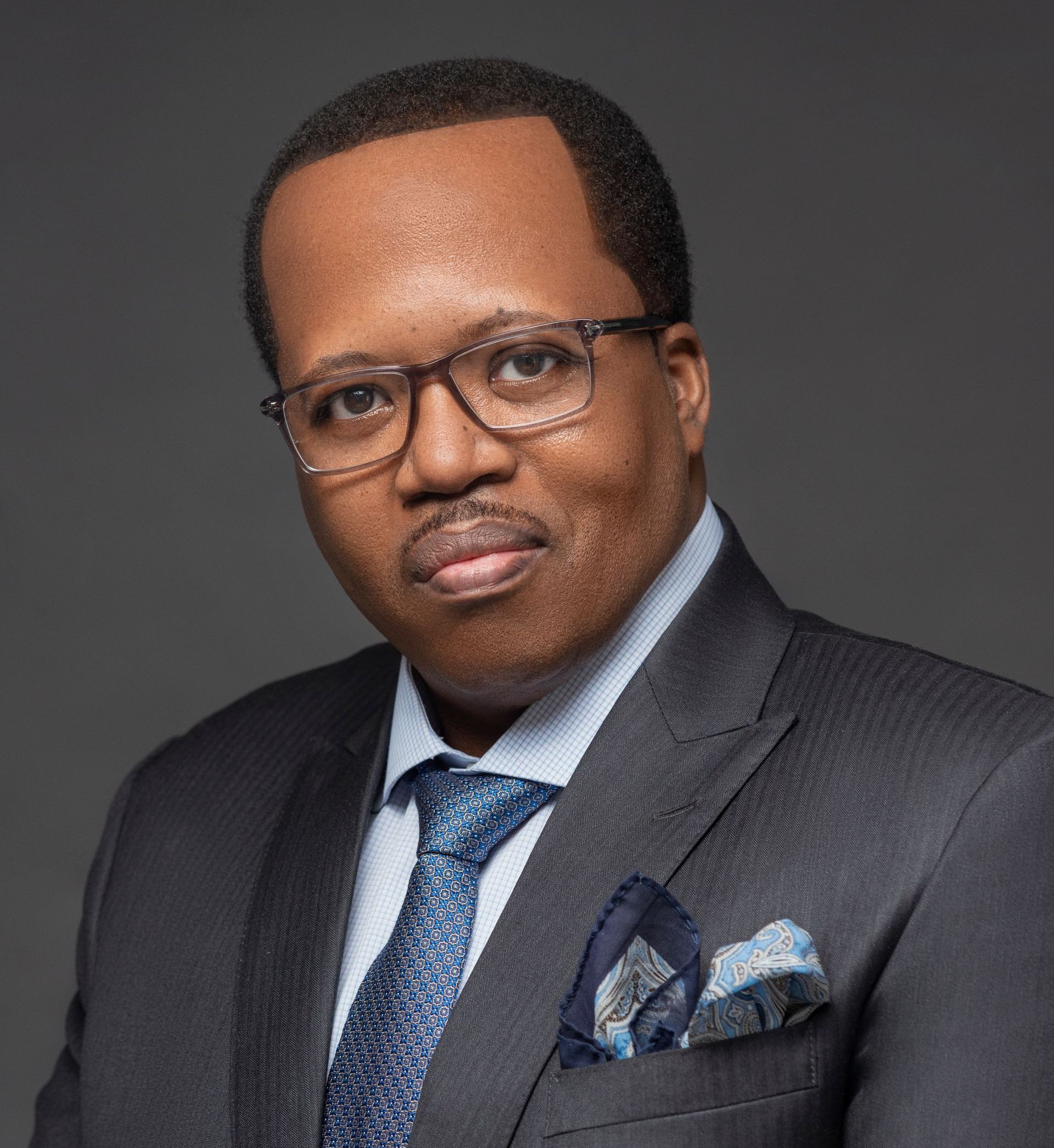 Man in suit and glasses looking at the camera, wearing a patterned tie and pocket square, with a gray backdrop.