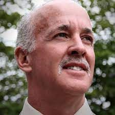 Bald man with graying hair and mustache, looking upwards. Outdoors with blurred green background.
