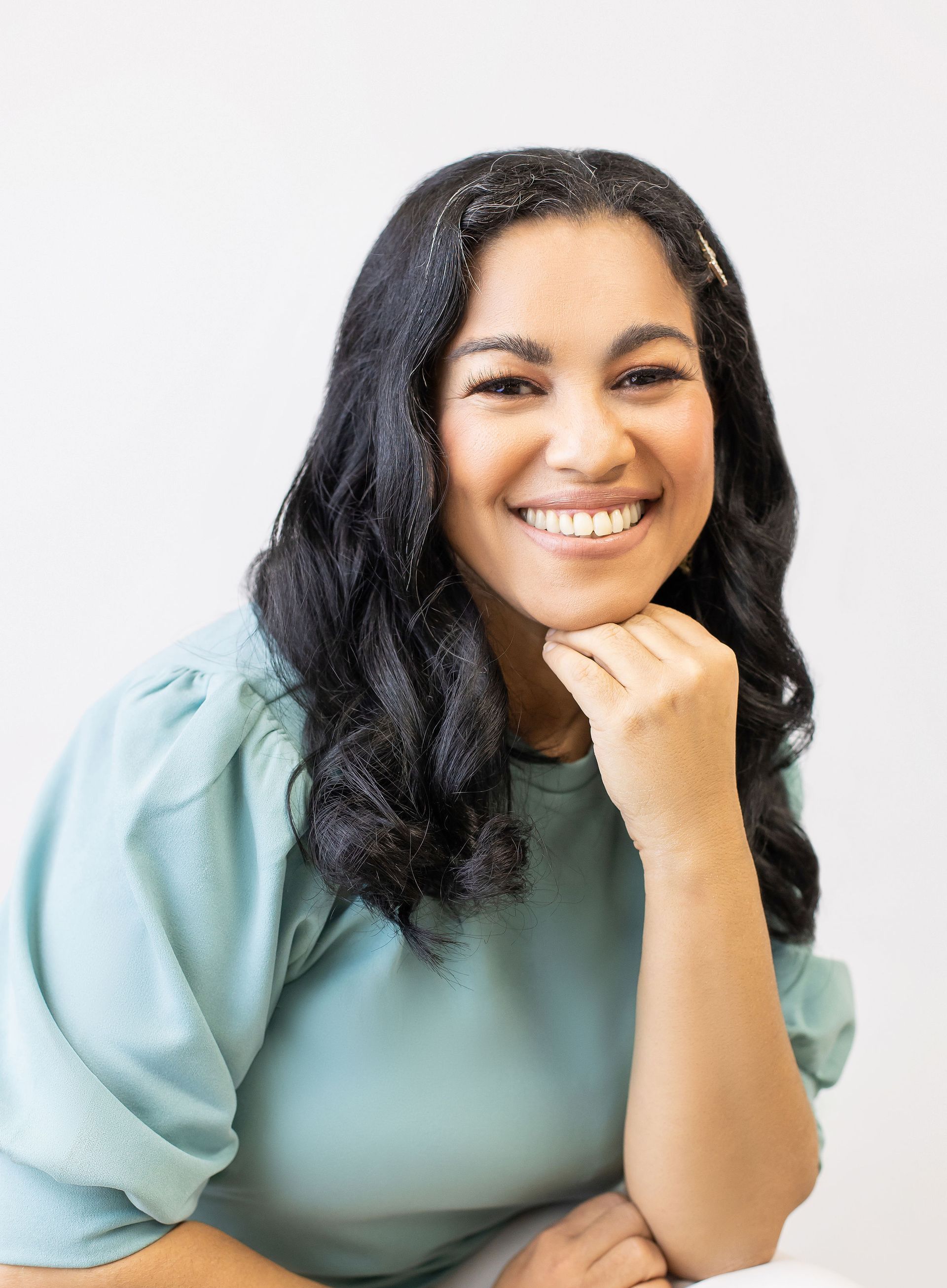 Woman smiling, leaning on hand. She wears a light blue top, dark hair, against a white backdrop.