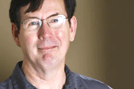 Man wearing glasses smiles, headshot against neutral background.