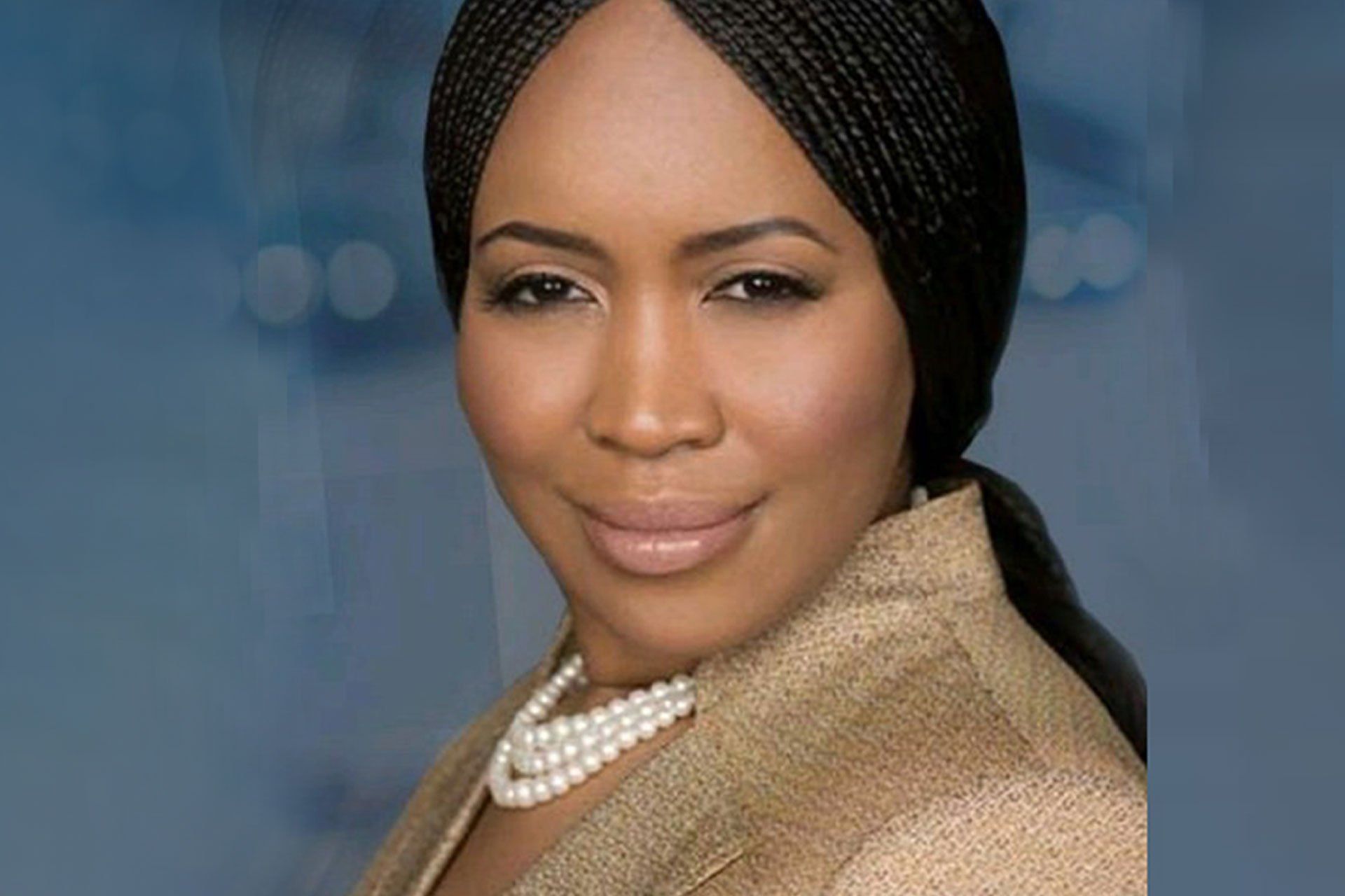 Woman with braided hair, wearing a gold jacket and pearl necklace, smiles against a blurred blue background.