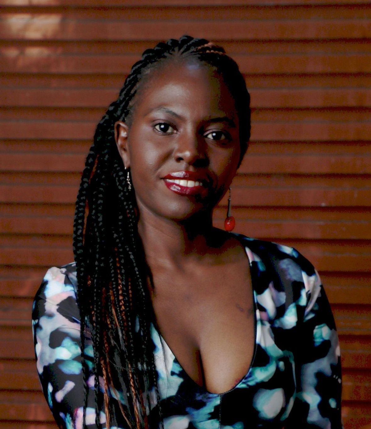 Woman with braided hair smiling, wearing a patterned top, in front of wood panels.