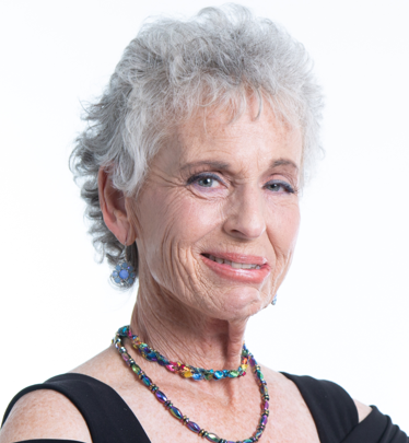Woman with short, gray hair smiles, wearing a black top and a colorful beaded necklace.