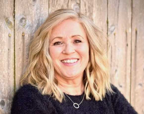 Blonde woman smiling, wearing a black sweater and silver necklace, against a wooden background.
