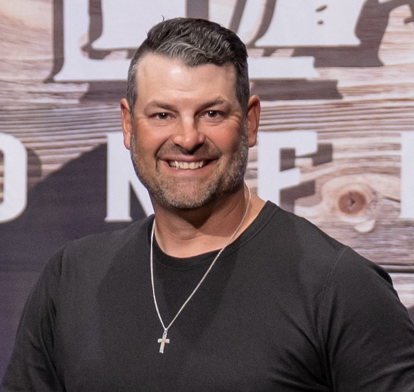 Man smiling, wearing black shirt and cross necklace, in front of wooden background.