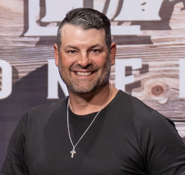 Man smiling, wearing black shirt and cross necklace, in front of wooden background.