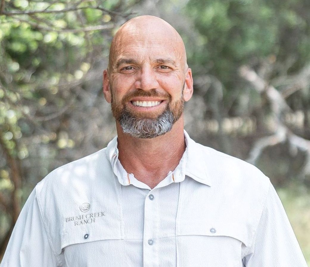 Bald man with a grey beard smiles, wearing a light blue collared shirt outdoors.