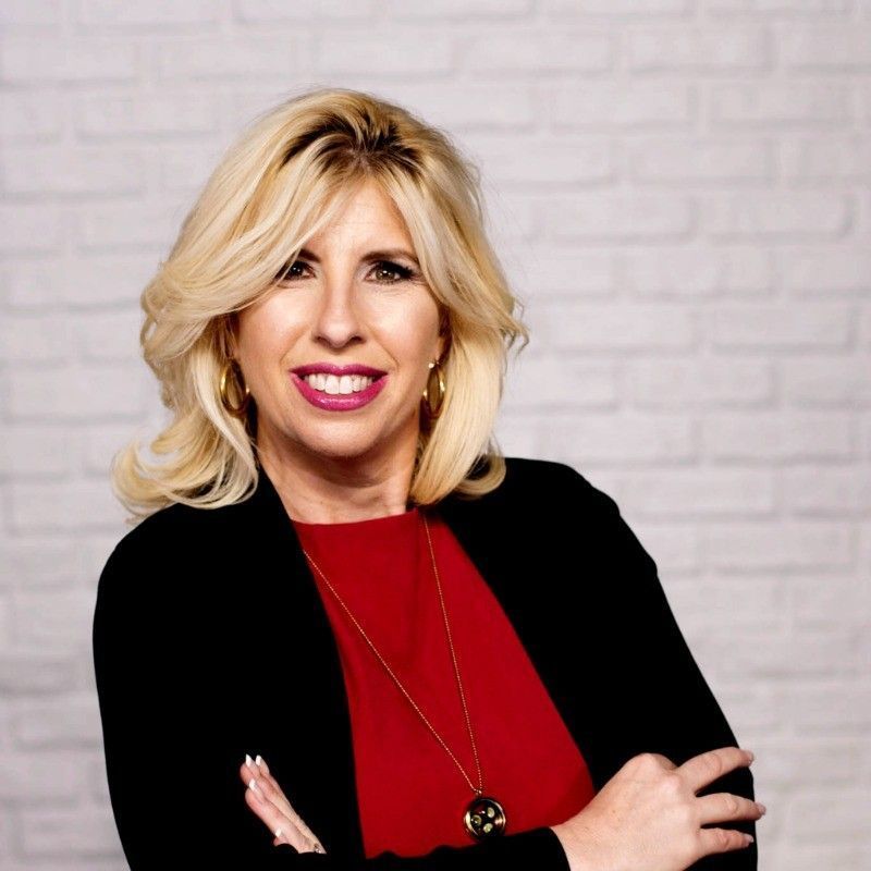 Woman with blonde hair, red top, black jacket, and gold necklace against white brick wall.