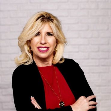 Woman with blonde hair, red top, black jacket, and gold necklace against white brick wall.