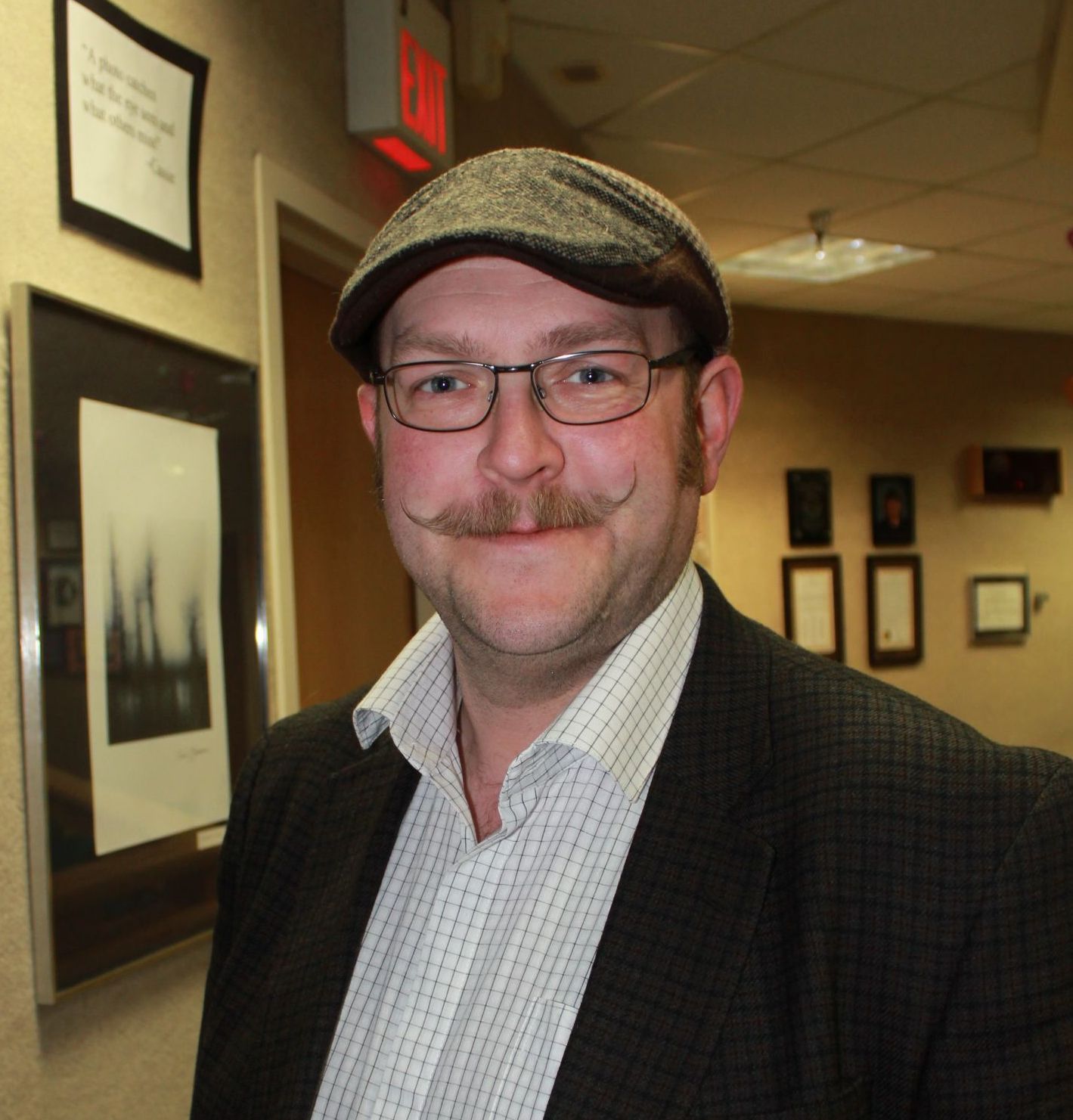 Man with glasses and mustache wearing a tweed cap and blazer in a hallway.