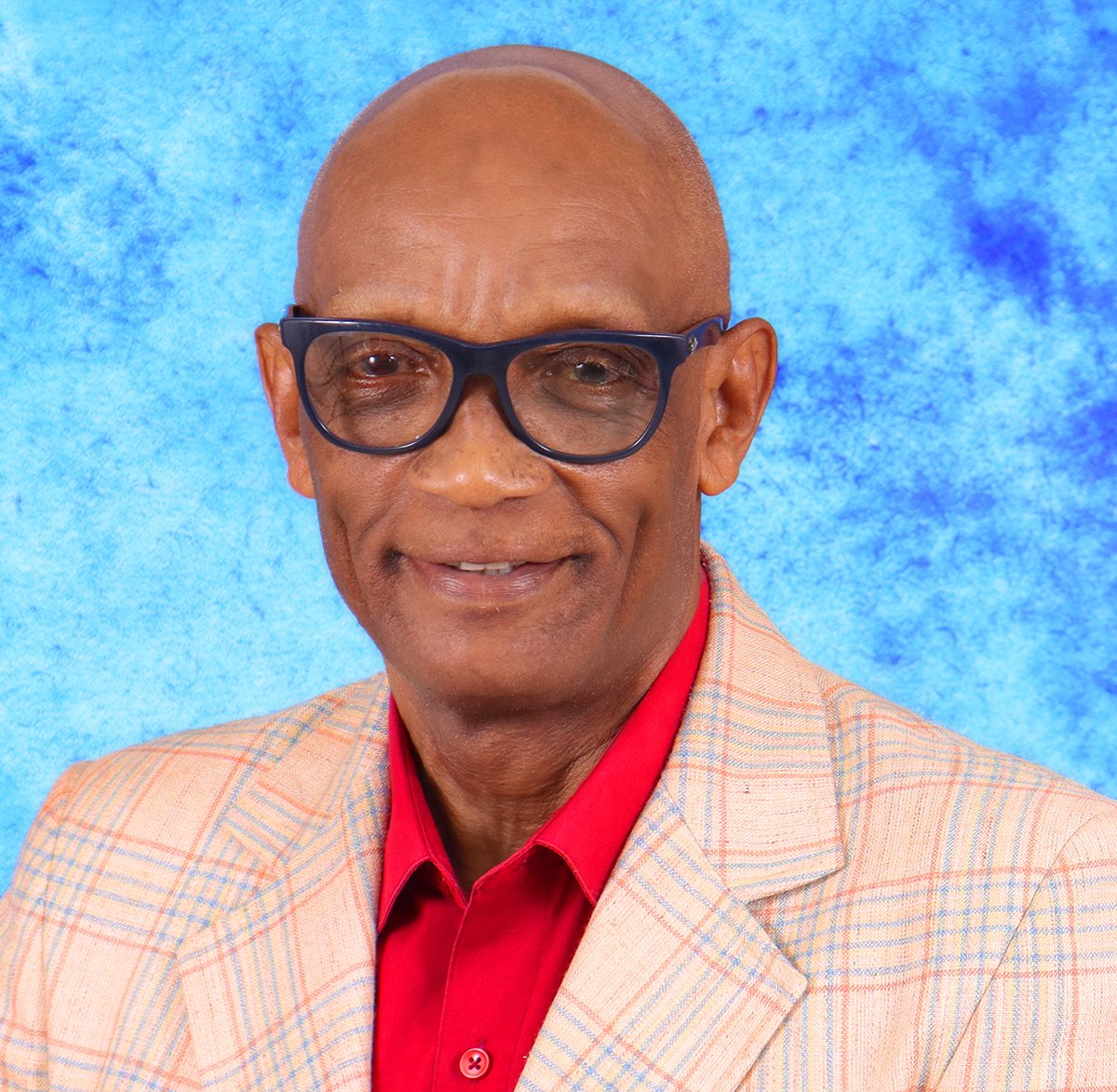 Man wearing glasses, red shirt, and plaid blazer smiles in front of a blue background.