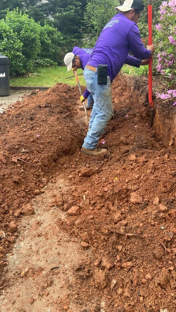 Two workers in purple shirts dig a trench in a yard, one holding a level rod, the other using a tool.
