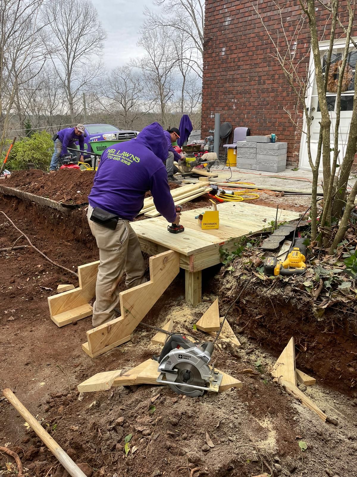 Construction of a wooden deck with stairs outdoors; workers in purple shirts, tools, and exposed dirt.