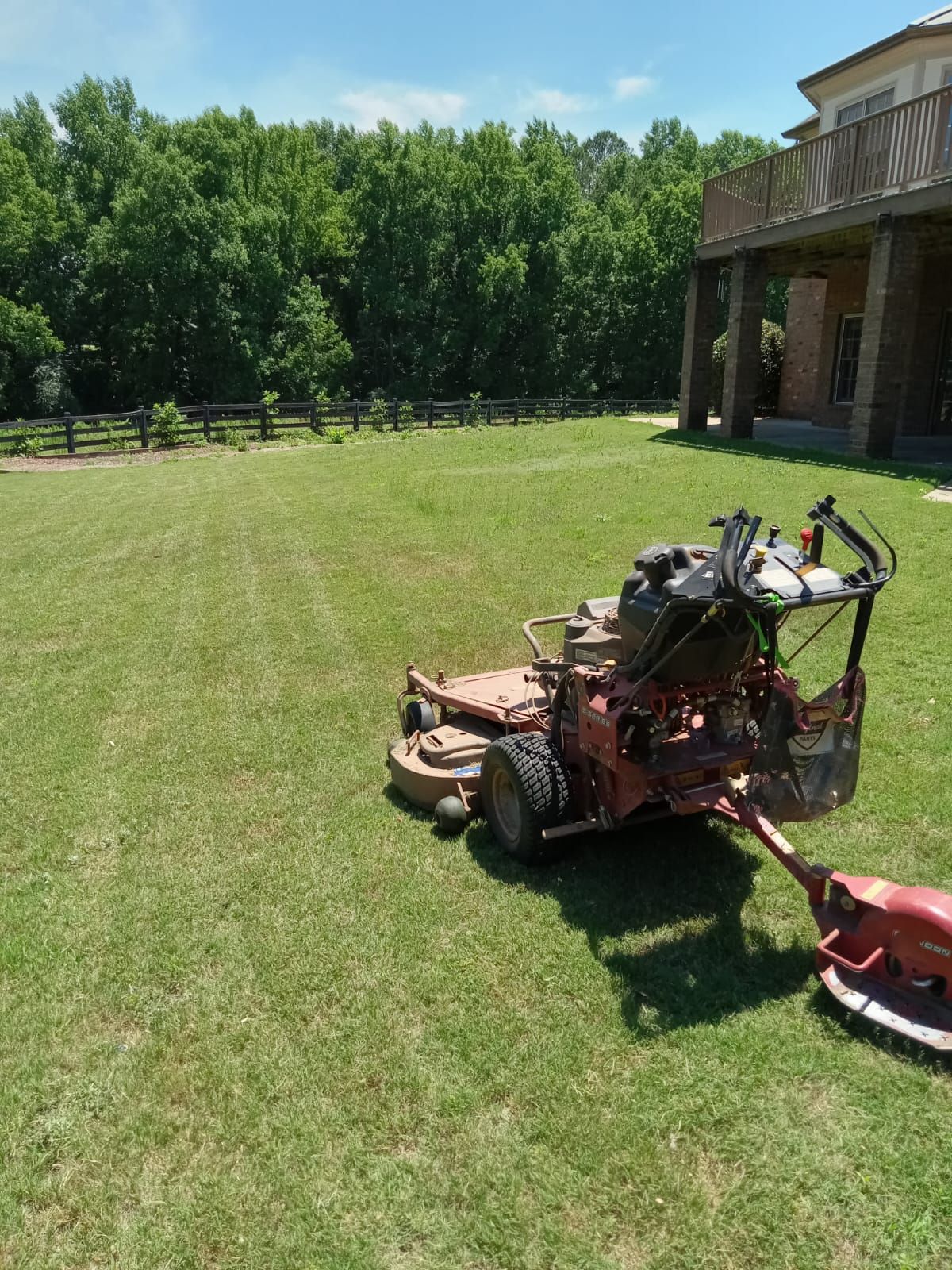 Lawnmower on a green lawn near a house, trees, and fence on a sunny day.