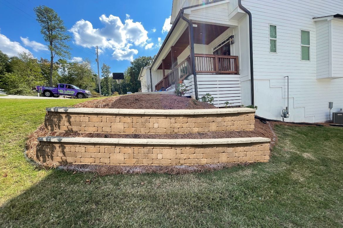 Terraced brick retaining wall with mulch and steps, in front of a white house with a porch.