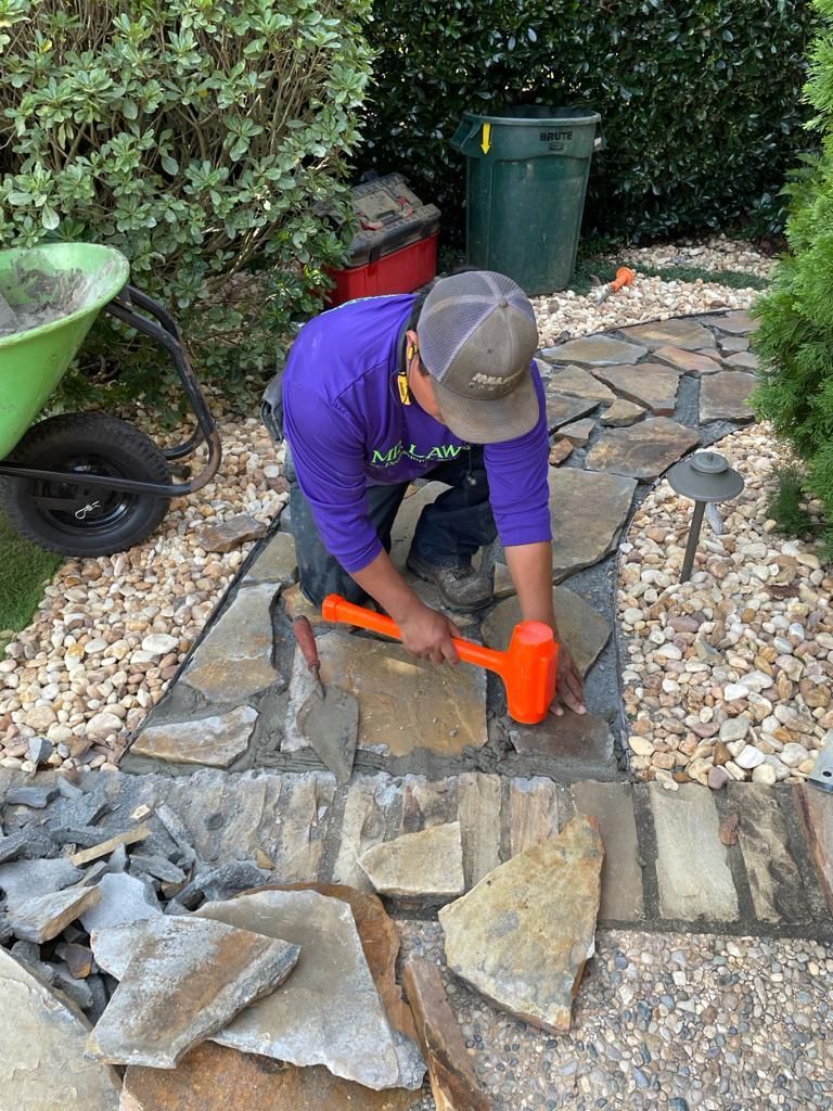 Man using a mallet to lay stone pathway. Outdoors, green wheelbarrow and trash can in background.