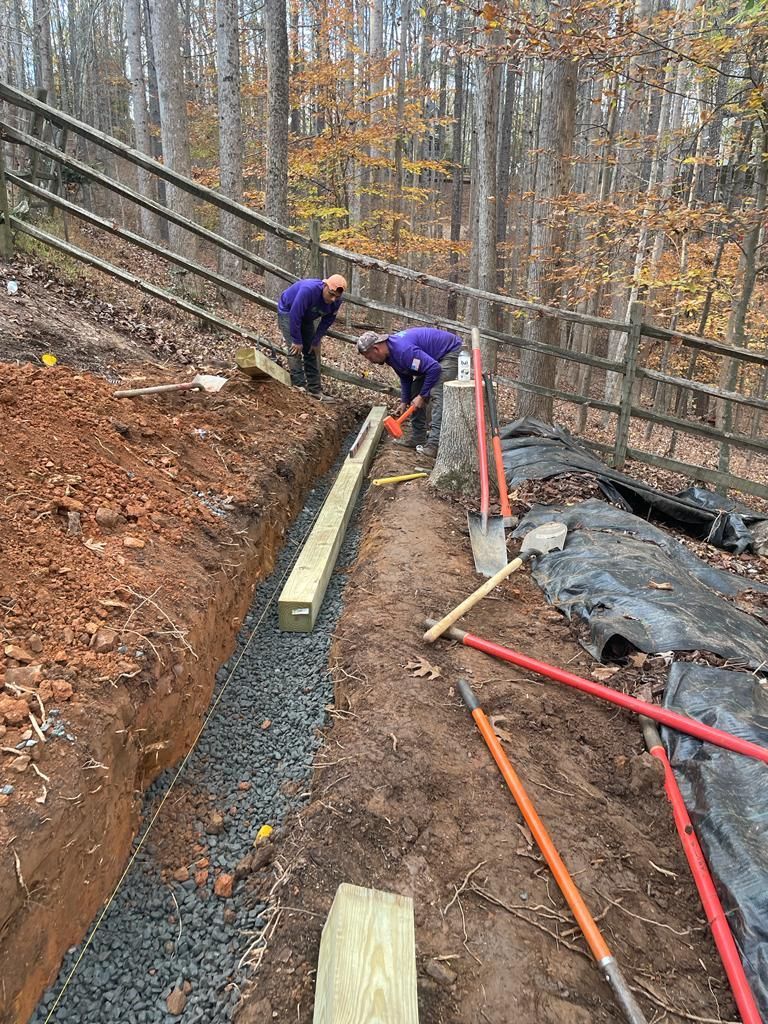 Two people in purple working on a trench filled with gravel; fence and trees in background.