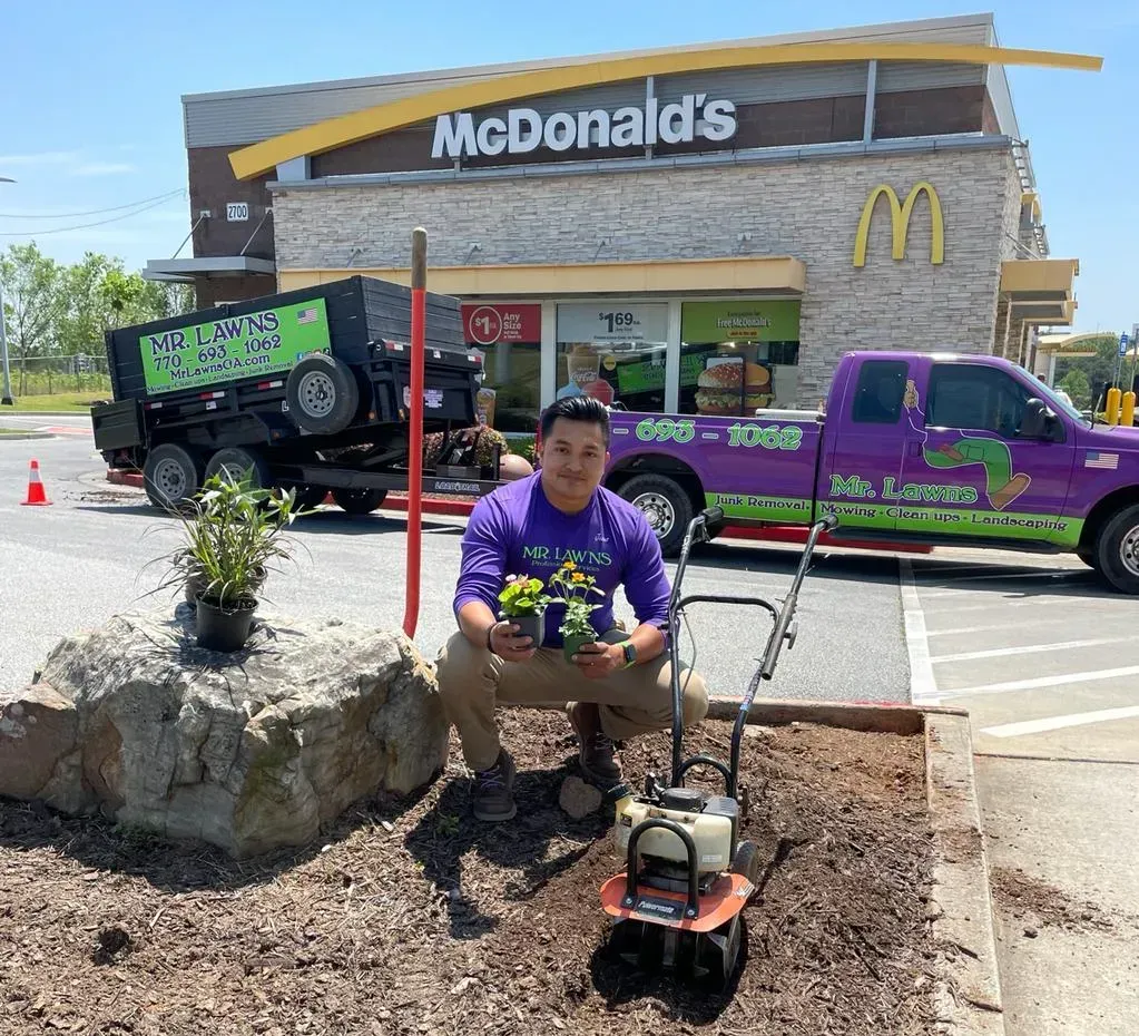 Man kneeling with plants, rototiller in front of McDonald's, purple landscaping truck and trailer in the background.