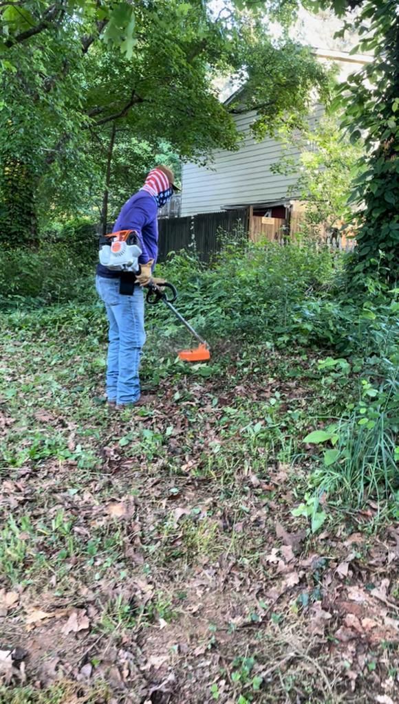 Person using a string trimmer in a grassy area with a wooded background.