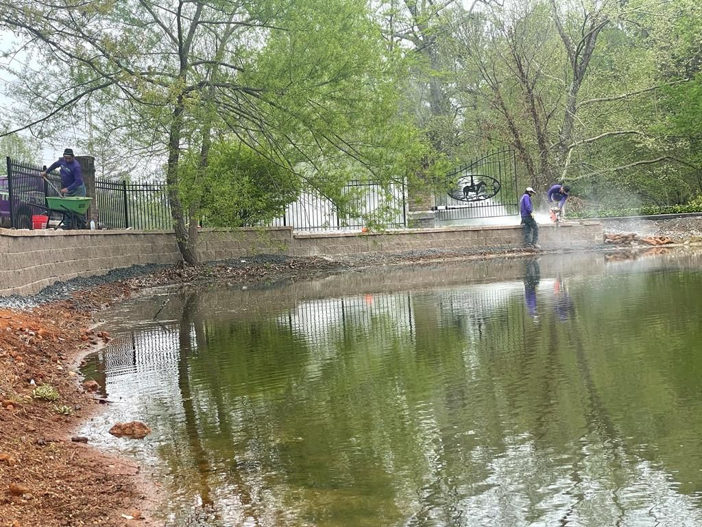 A pond with workers cleaning debris. Trees surround the water. A black fence sits behind the workers and pond.