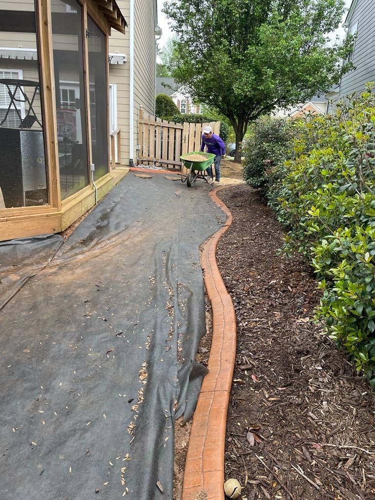 Person using a wheelbarrow in a backyard. A black fabric covers the ground. Decorative border and bushes line the path.